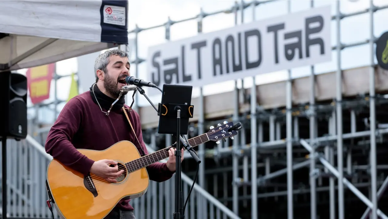 Man with beard singing into microphone whilst playing acoustic guitar at outdoor venue.