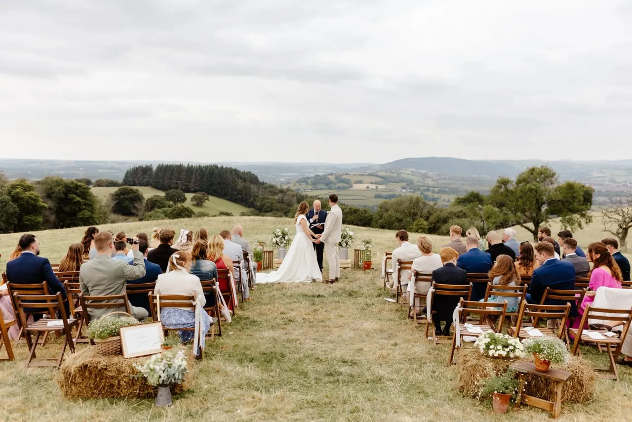 Outdoor wedding ceremony in a field with guests seated on wooden chairs facing the bride and groom, rolling countryside landscape in the background.