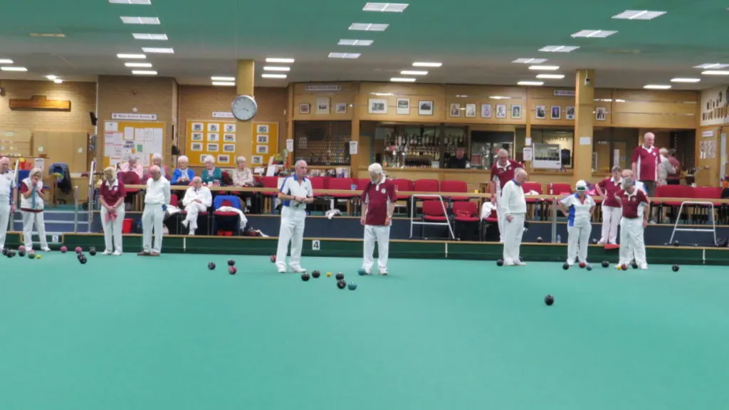 Members of various ages playing bowls indoors on a turquoise green, with spectators watching from seating behind.