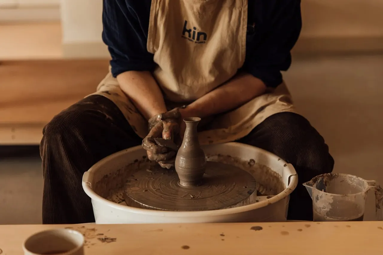 A person at a pottery wheel shapes clay into a small vessel whilst seated at the wheel.