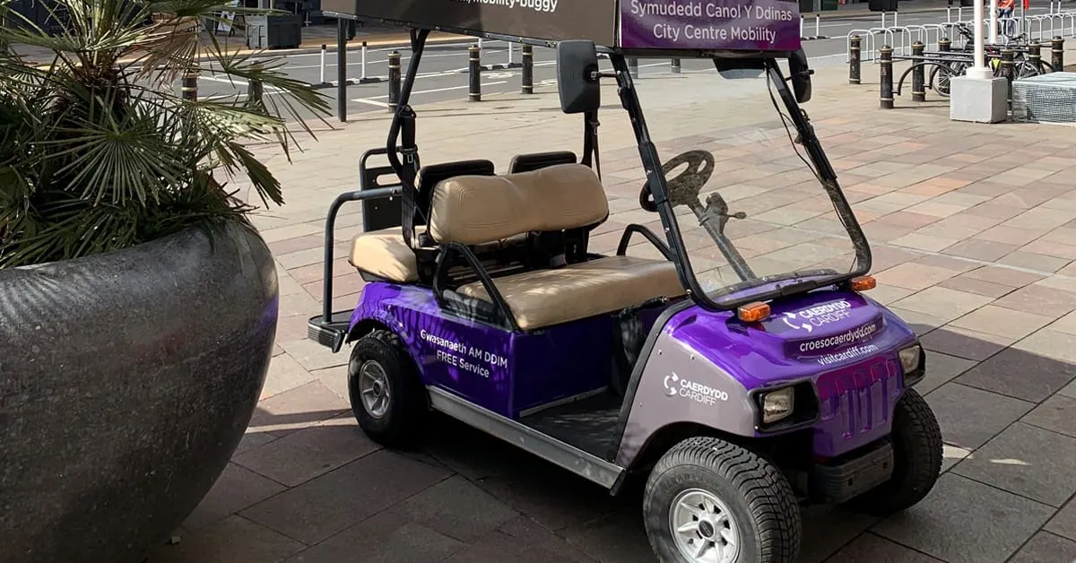 Purple and white golf buggy parked on a city centre paved area with planters and signage visible.