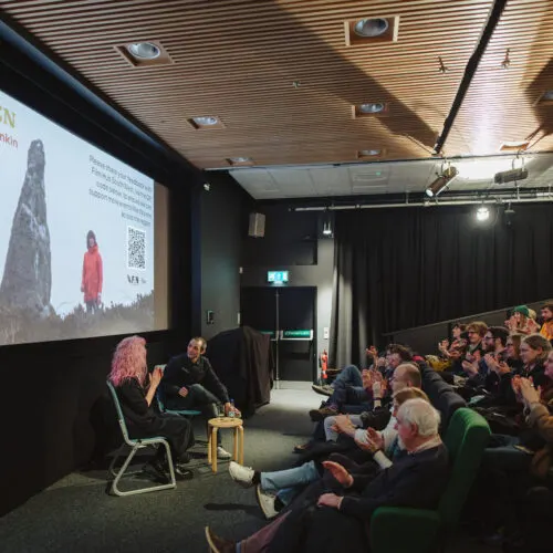 Audience seated in a cinema watching a projected presentation with two speakers on stage.