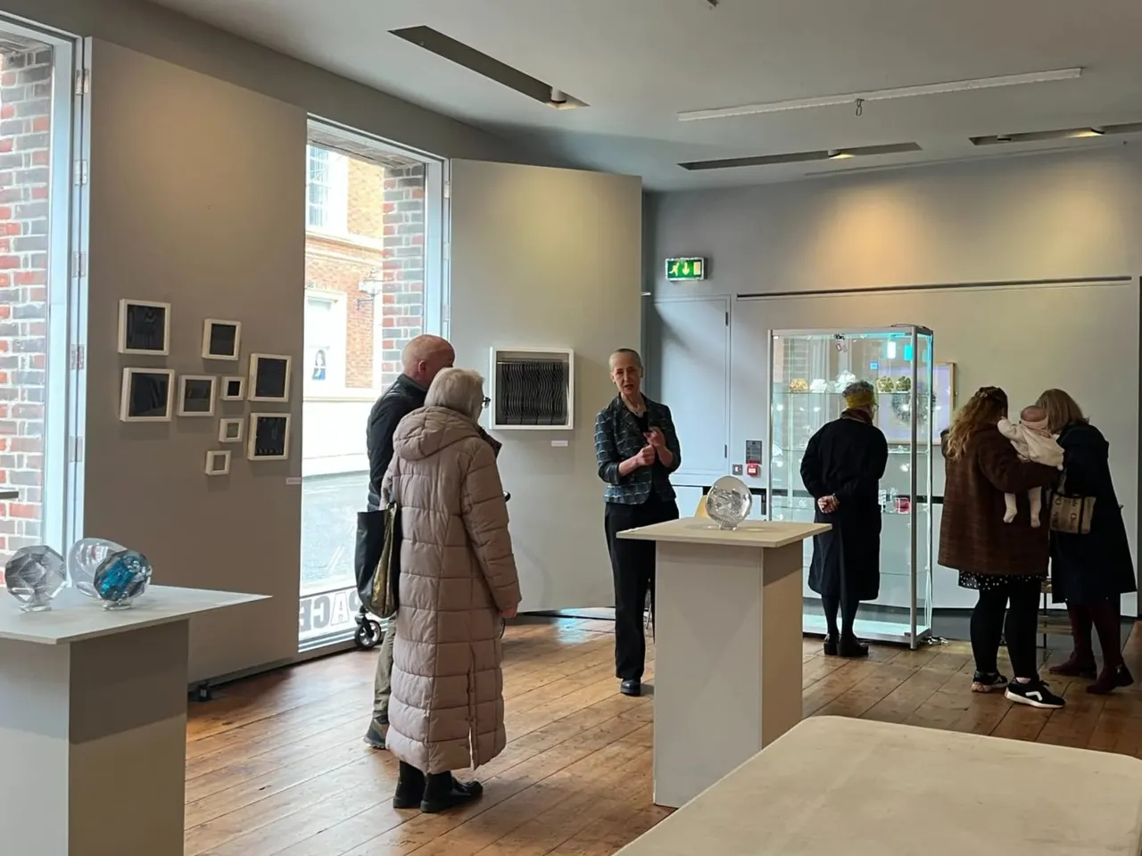 Visitors browse artwork in a contemporary gallery space with display cases, wall-mounted framed pieces, and a guide speaking to guests.