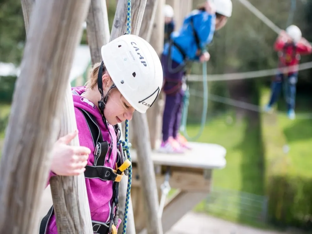Child in white helmet and purple harness climbing wooden pole at outdoor adventure activity course.