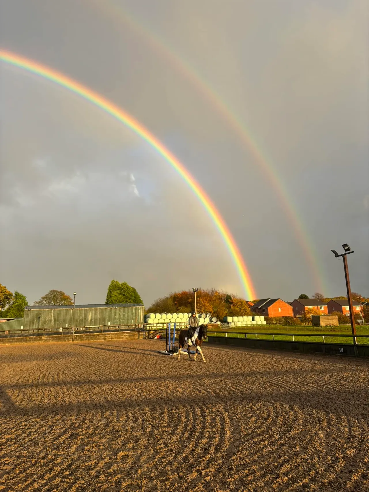 A double rainbow arcs over a rider on horseback in a sand arena, with farm buildings and hay bales visible in the background.