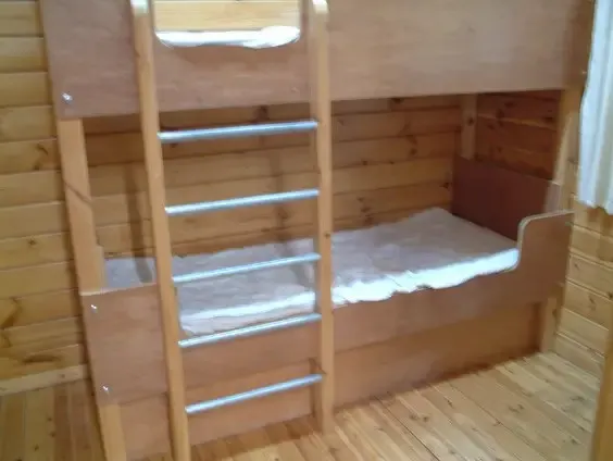 Wooden bunk bed with ladder inside a wooden cabin, lit by natural light from above.