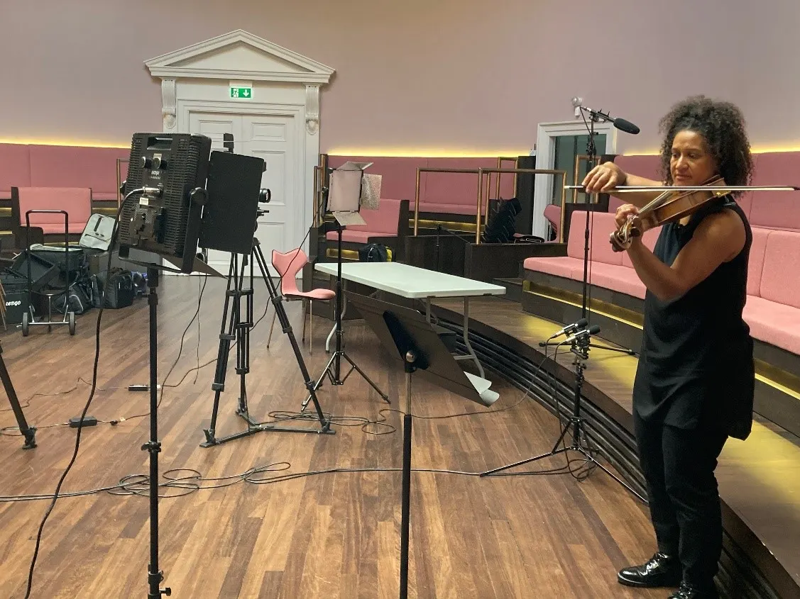 Musician playing violin in a concert hall with professional recording equipment and pink seating visible in the background.