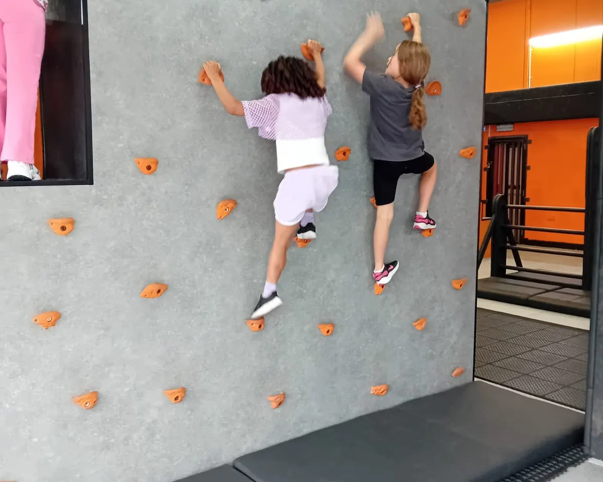 Two children climbing an indoor rock wall with orange hand and foot holds.