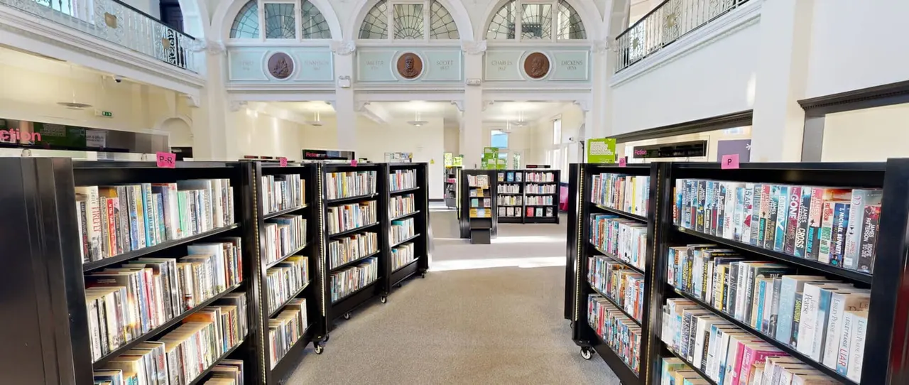 Interior of a public library with black shelving units displaying DVDs and books, arched windows and period medallions on upper walls.