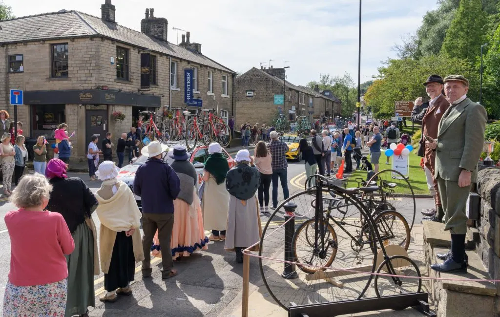 Outdoor community event in a town street with vintage bicycles on display, crowds of visitors, historic stone buildings, and people in period costume.