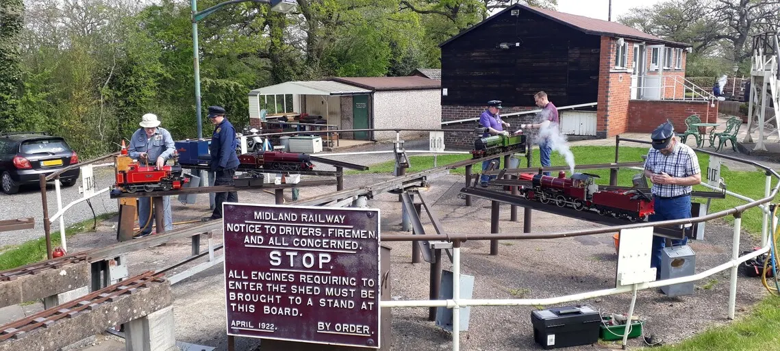 Men operating miniature steam locomotives on a gauge railway track in an outdoor yard with sheds and trees.