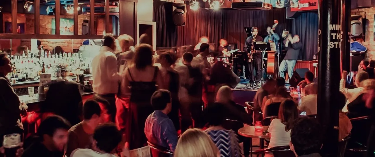 Crowded jazz bar with live band performing on stage whilst patrons dance and socialise under red lighting.