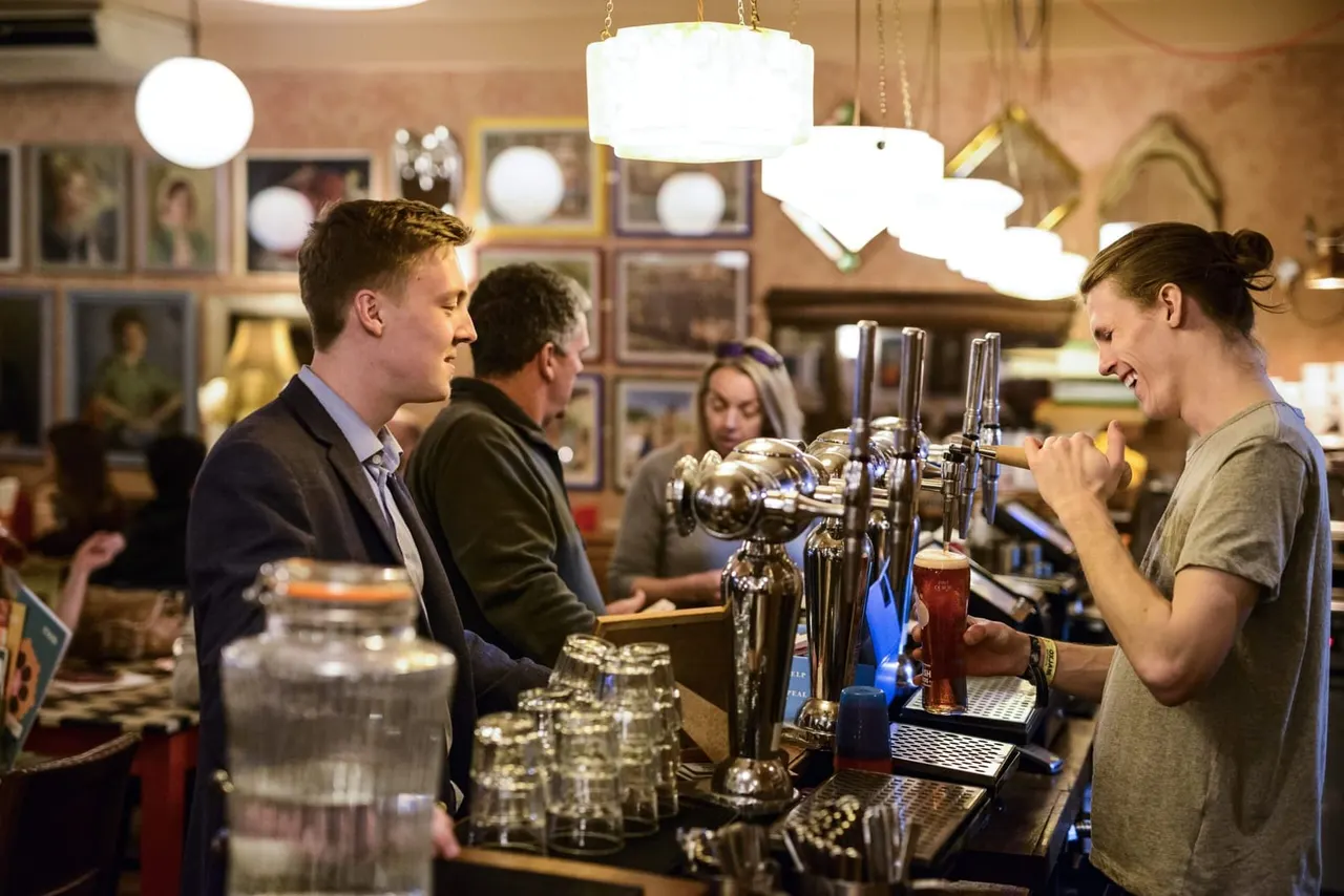 Bartender pouring a drink for a customer at a pub bar with other patrons in the background.
