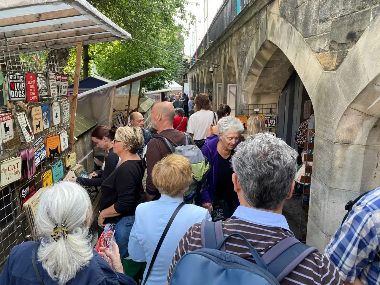 Crowd of visitors browsing market stalls beneath a stone bridge arch in an urban alleyway.