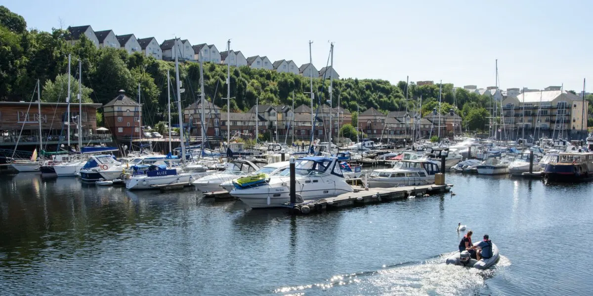 Two people in a dinghy on the water at a busy marina with moored sailboats and waterfront buildings.