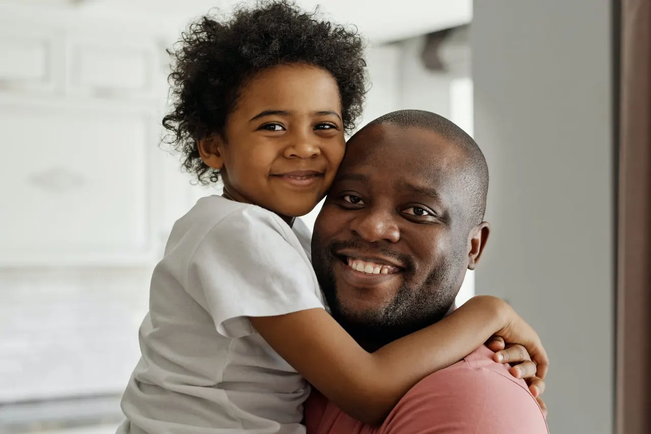 A young child hugs an adult man from behind whilst both smile at the camera in a bright indoor setting.