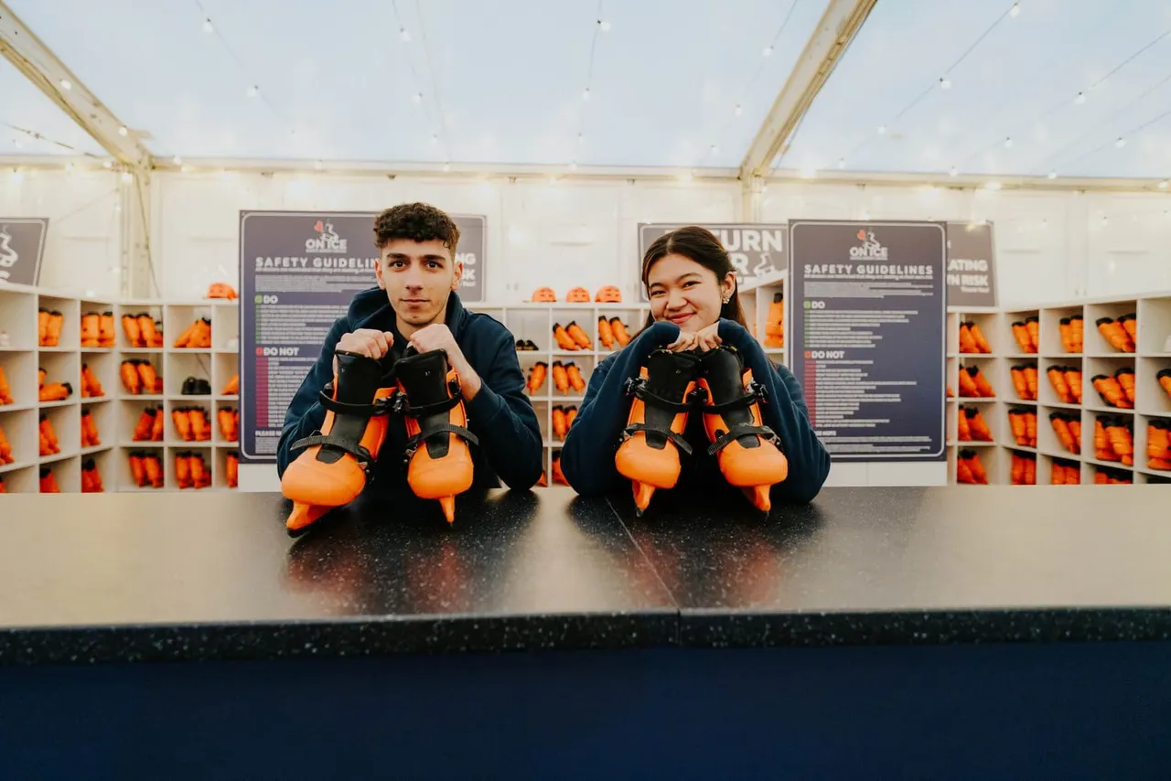 Two young people holding orange and black ice skates at a counter inside an ice skating venue, with safety guidelines posters and equipment shelves visible behind them.