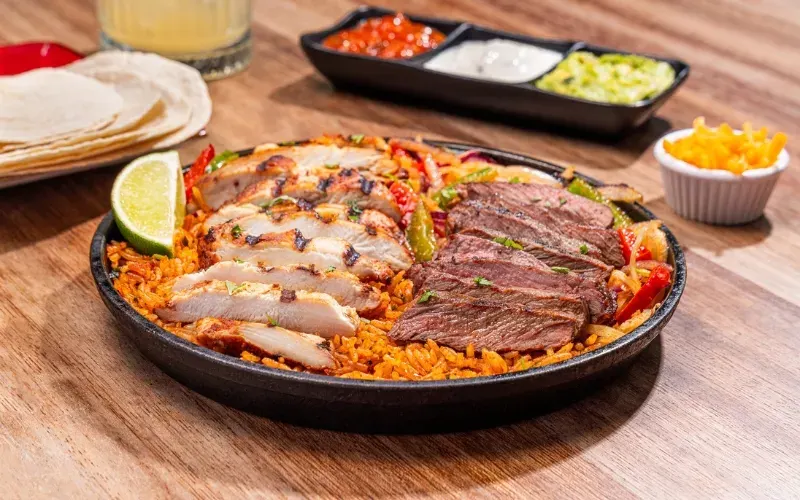 Mixed grilled meats and seafood sizzling on a cast-iron plate with rice and peppers, surrounded by condiments and tortillas.