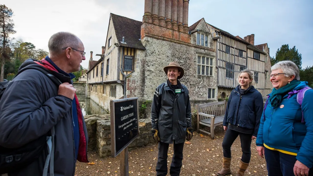 A National Trust guide leads a group of visitors on a garden tour outside a historic half-timbered medieval manor house.
