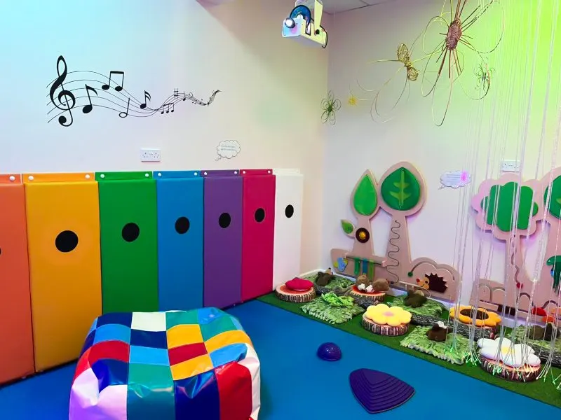 Colourful play room with rainbow storage boxes, sensory play equipment, musical notes on wall, and nature-themed decorations.