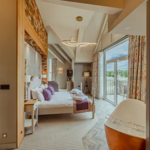 Modern bedroom with double bed, purple cushions, exposed brick wall, and access to a balcony overlooking countryside.