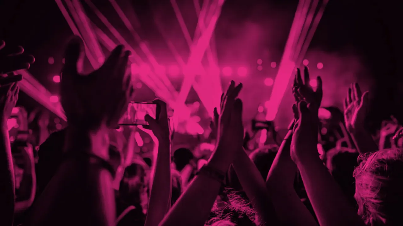 Crowd of people dancing and cheering at a concert with bright pink stage lighting and laser effects overhead.