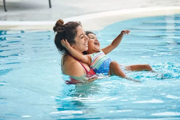 Woman and young child swimming together in a pool, child reaching out while being supported.
