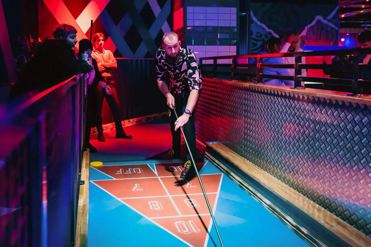 Man playing shuffleboard at an indoor entertainment venue with neon lighting and spectators watching from behind barriers.