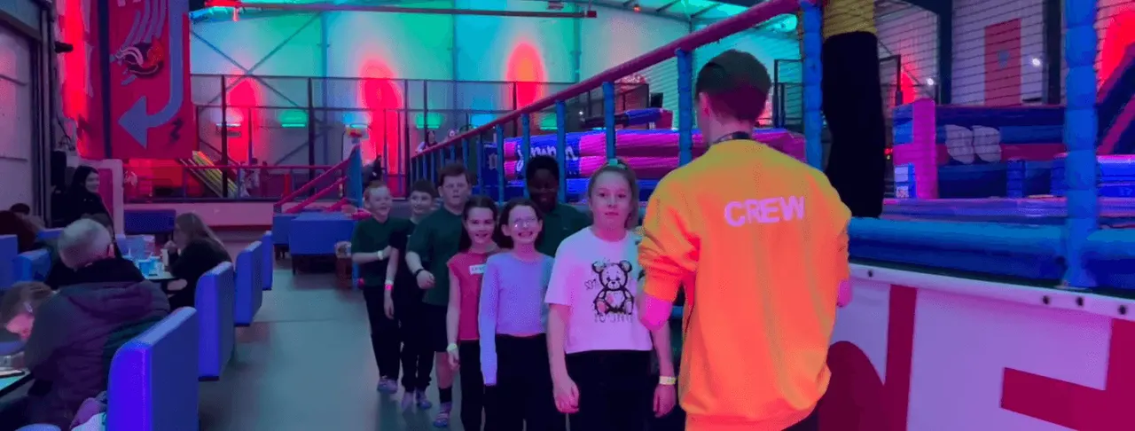 PHOTOGRAPH

Group of children and staff member in neon-lit indoor trampoline park with colourful lighting and padded structures.
