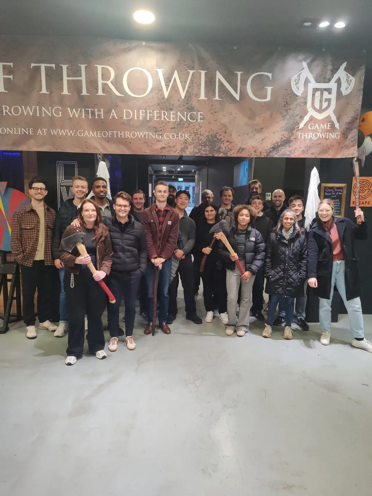 A group of about twenty-five people posing together indoors at an axe throwing venue, many holding axes.