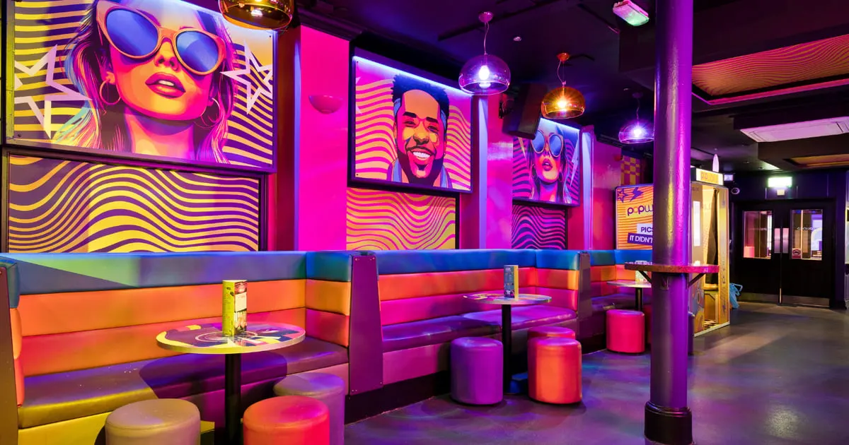 A brightly lit nightclub interior with vibrant neon purple, pink, and yellow lighting, colourful booth seating, and digital screens displaying pop art portraits on the walls.