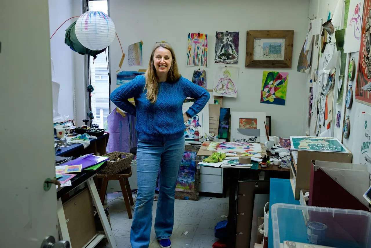 Woman in blue jumper standing in art studio surrounded by colourful artwork, sketches and creative materials on walls and tables.