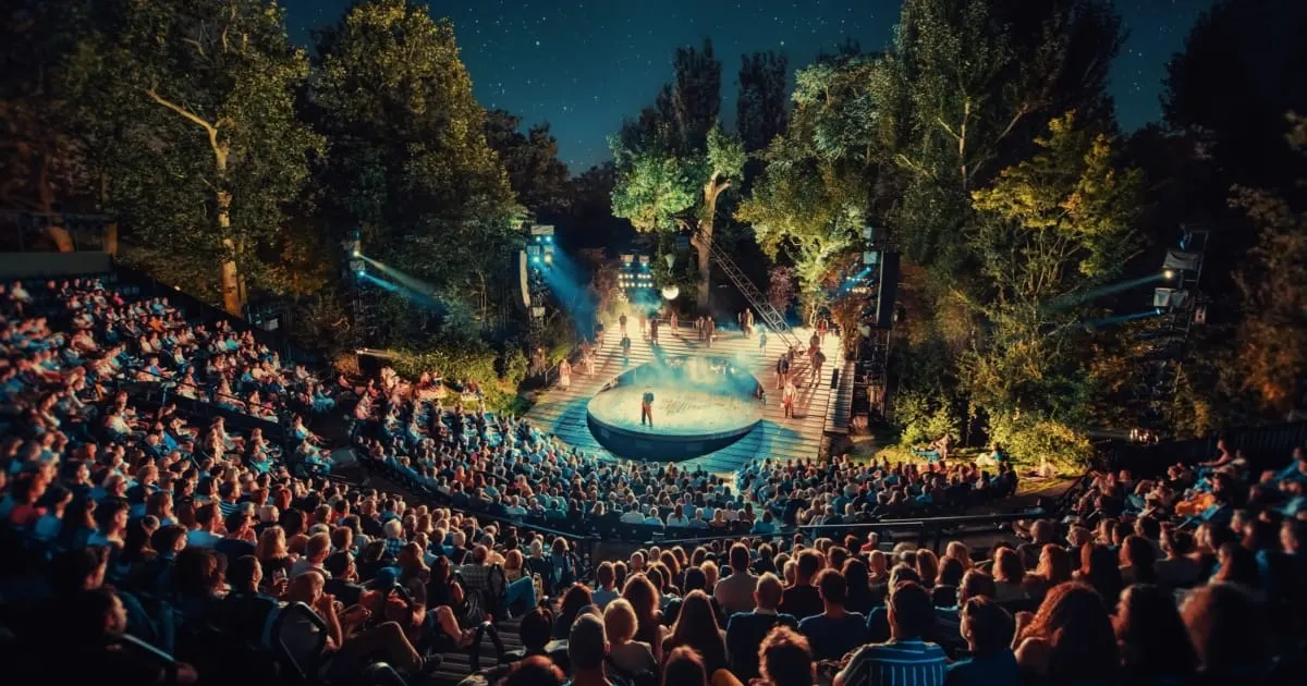 Large outdoor amphitheatre at night with hundreds of seated spectators watching a theatrical performance on an illuminated central stage surrounded by trees.