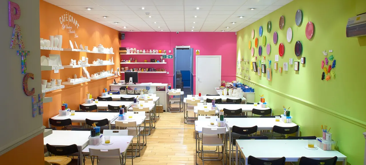 Bright, colourful pottery painting studio with multiple white tables, black chairs, shelves of blank ceramics, and decorative plates on vibrant orange, pink and green walls.