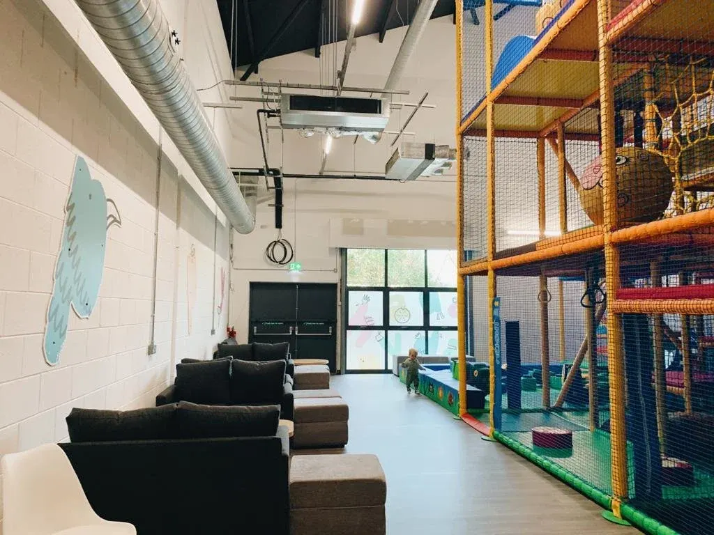 Indoor play area with seating area and large multi-level soft play structure with netting and climbing equipment.