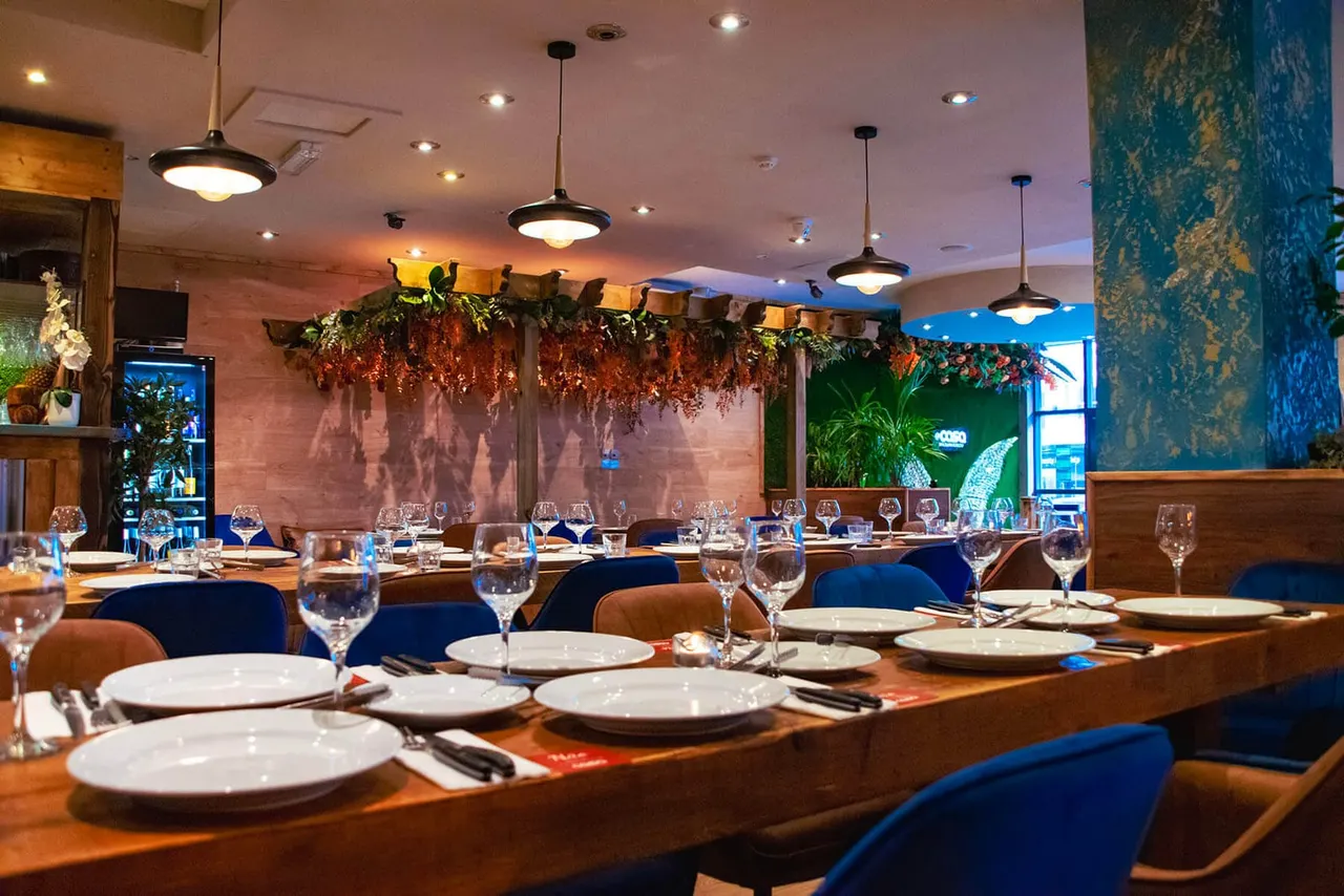 Modern dining room with long wooden table set for service, blue upholstered chairs, pendant lighting, and hanging floral arrangements overhead.
