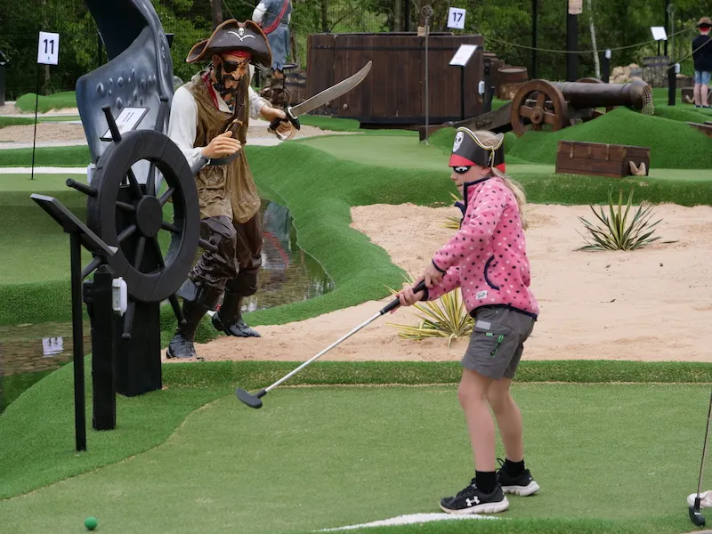 Child playing mini golf on a pirate-themed course with a costumed pirate character holding a ship's wheel.