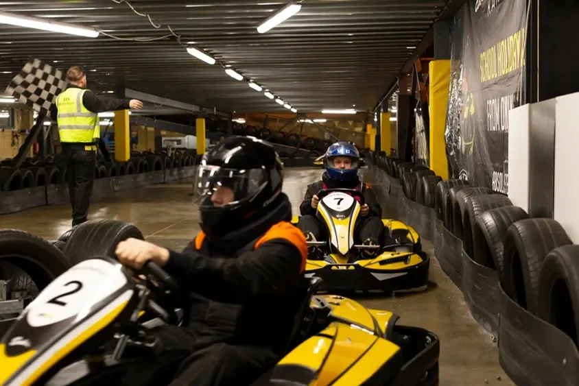 Indoor go-kart racing with multiple drivers in yellow and black karts on an asphalt track with barriers and staff.
