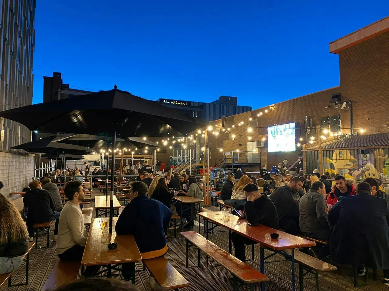 Outdoor rooftop terrace at dusk with crowds of people seated at long tables under black umbrellas and string lights, brick buildings surrounding the space.