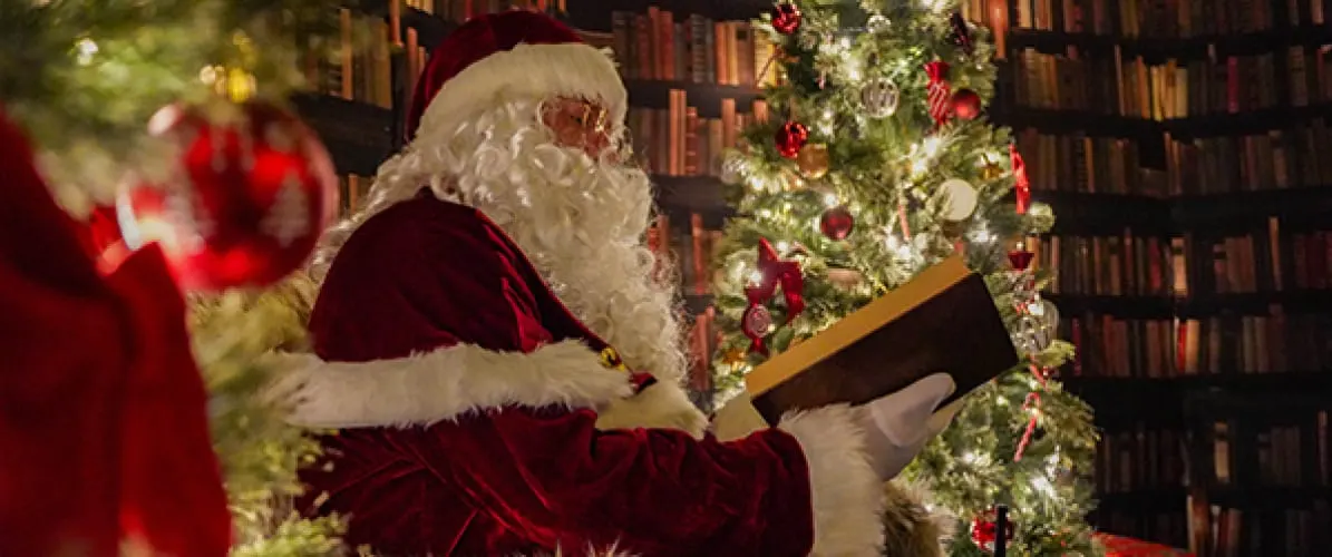Father Christmas reading a book in a festive library setting with decorated Christmas trees and baubles.
