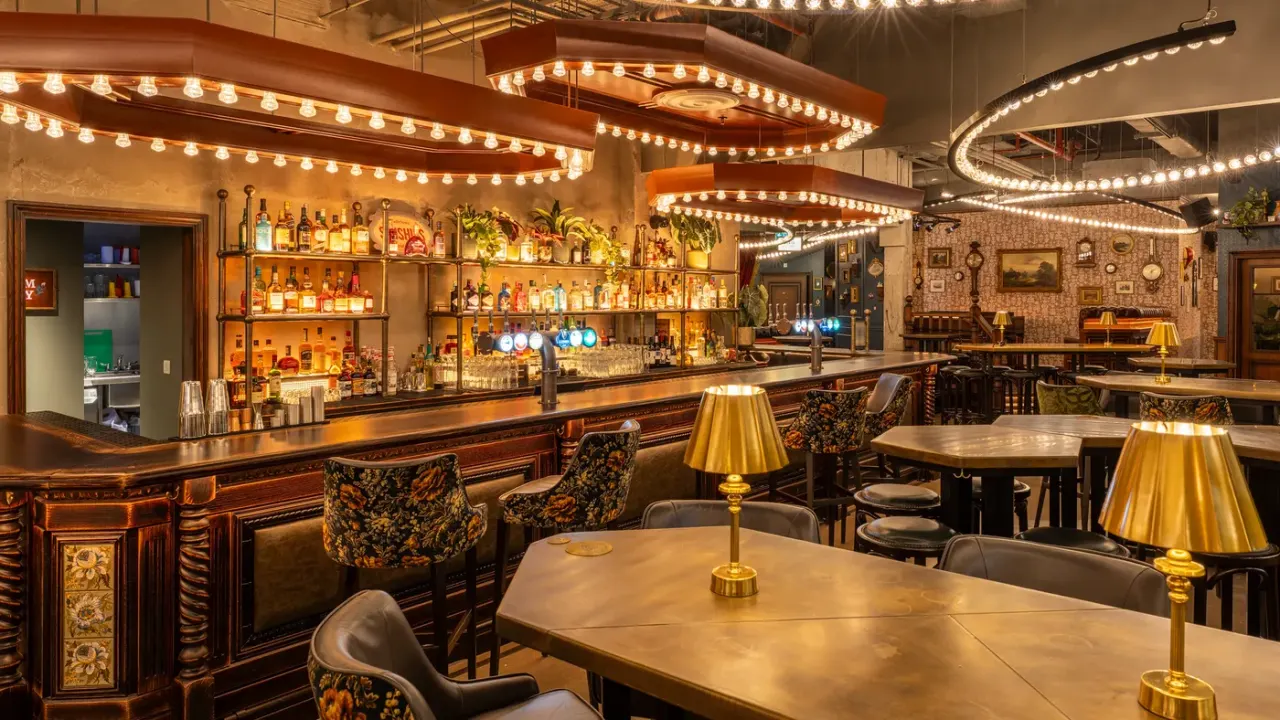 Upscale cocktail bar with illuminated shelving behind the counter, brass lighting fixtures, and vintage-style seating throughout the space.