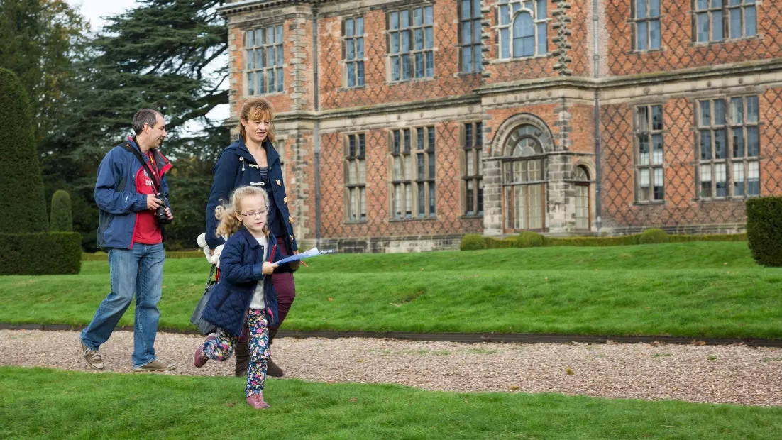 A family on a guided tour walks across the lawn in front of a large brick country house.