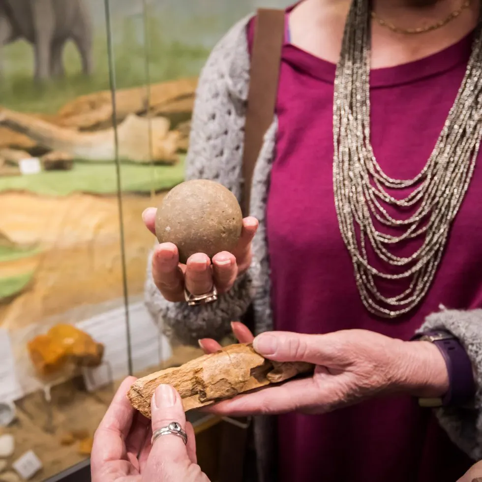A woman holds archaeological artefacts including a spherical stone object and pottery fragment, displayed indoors.