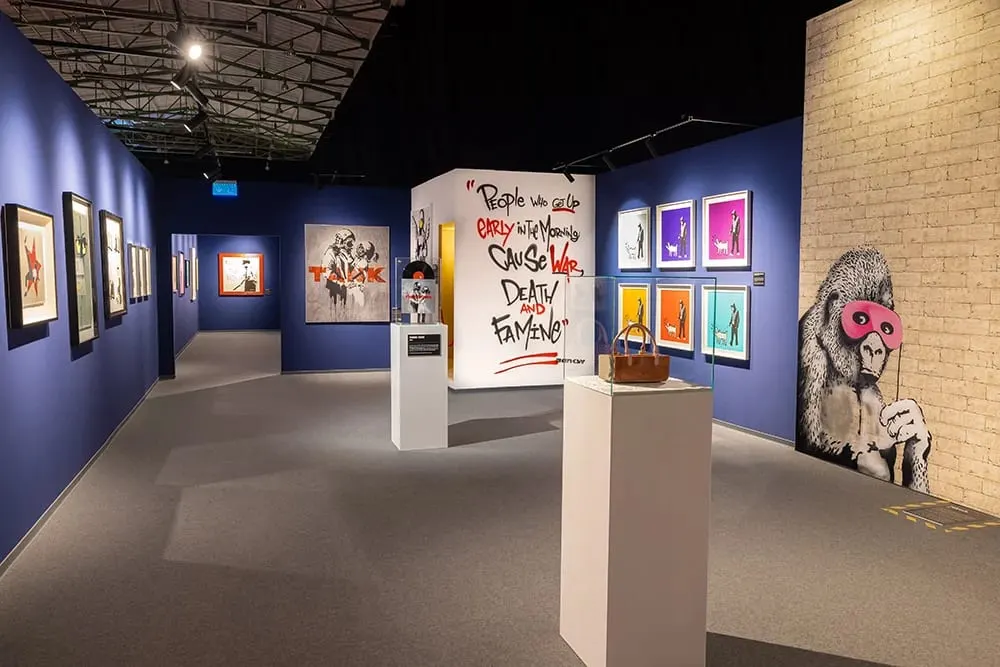 Contemporary art gallery interior with blue walls displaying framed prints and street art, including a large stencilled monkey figure holding a pink mask.