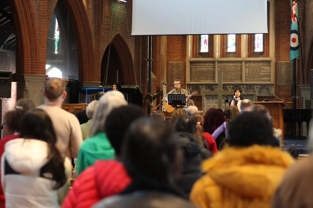 Audience seated in a brick church watching musicians perform on stage with a guitar and other instruments.