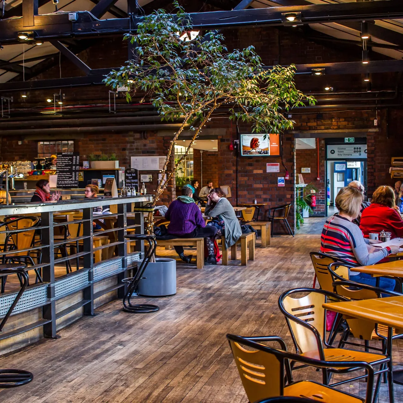 Interior of a modern café with visitors seated at tables, a central tree, counter service, and exposed brick walls with warm lighting.