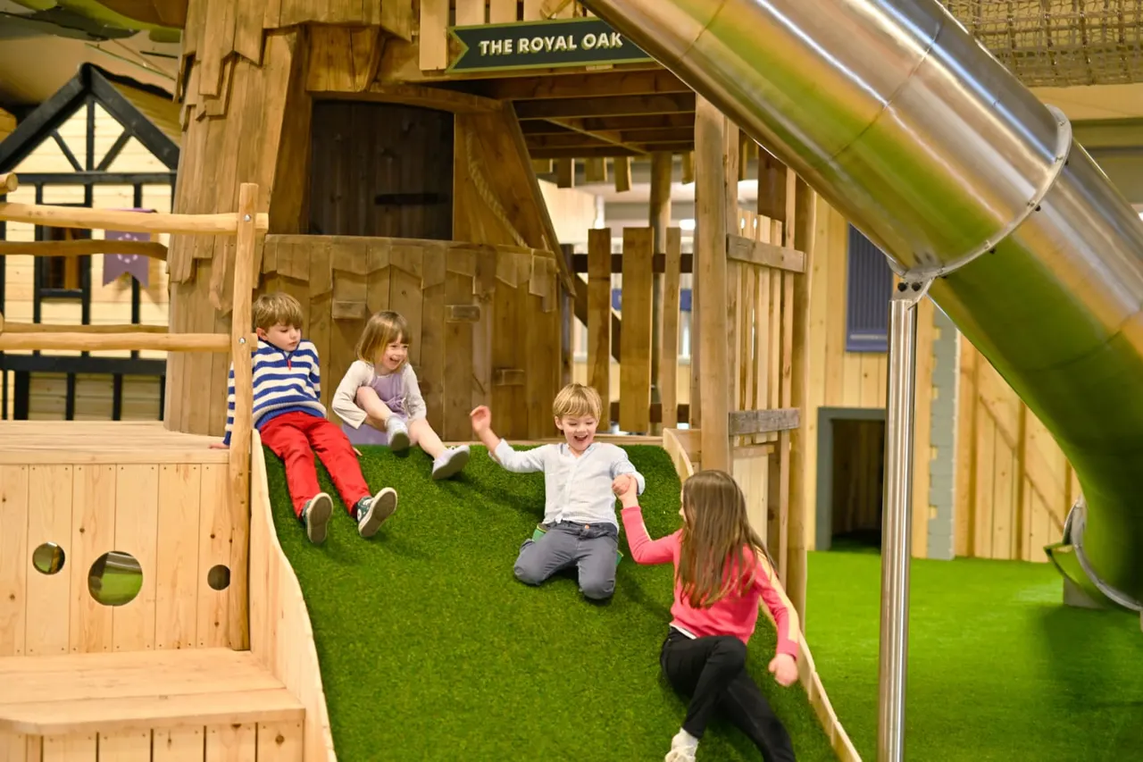 Four young children playing together on artificial grass inside a wooden playhouse structure with a slide.