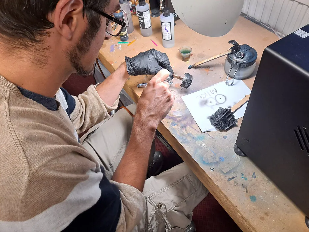 Man in glasses working on a miniature figure at a crafting table with paints and tools.