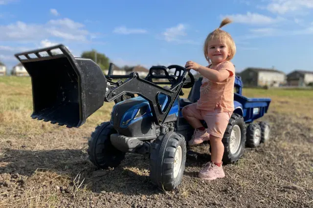 Young child standing next to a blue ride-on toy tractor with front loader bucket in a rural farm setting.