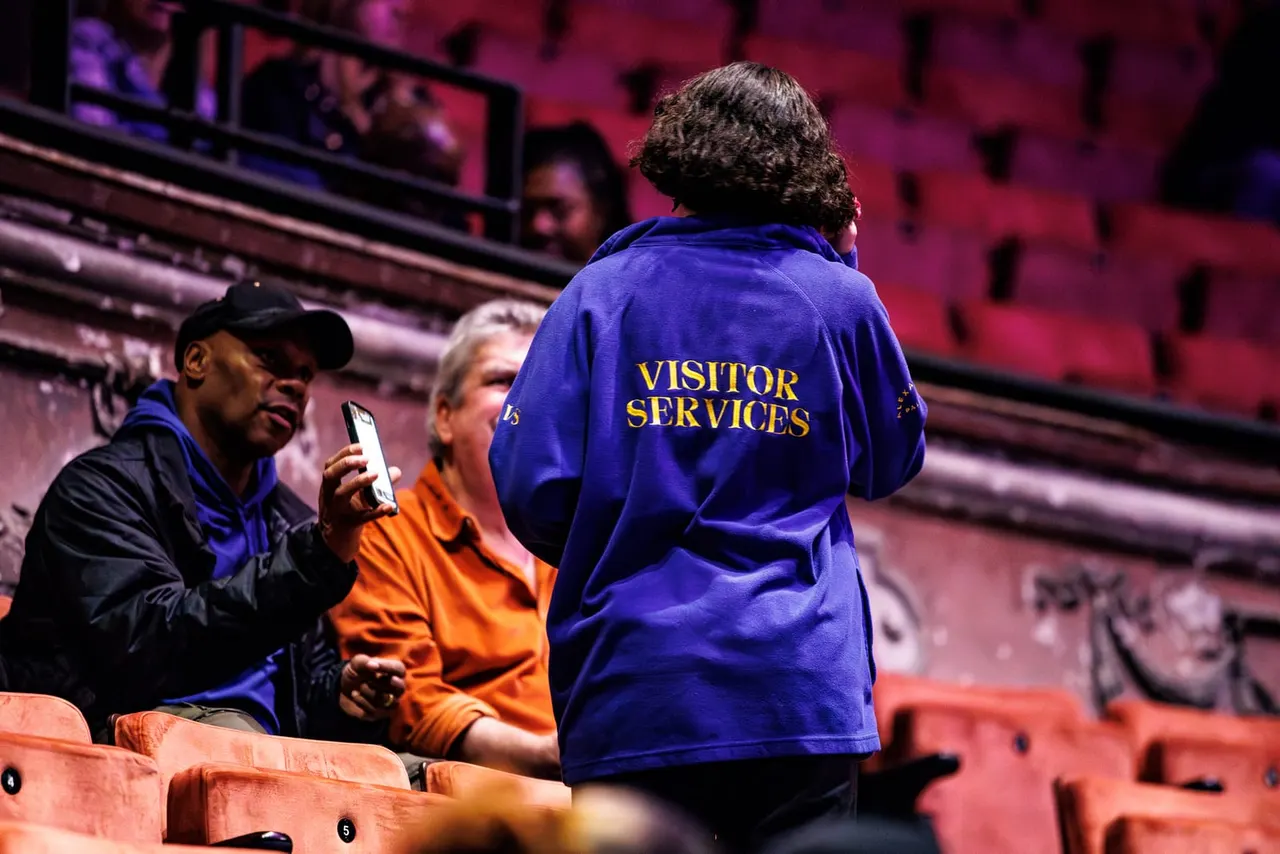 Visitor Services staff member in blue jacket assisting spectators in stadium seating with purple and pink lighting.